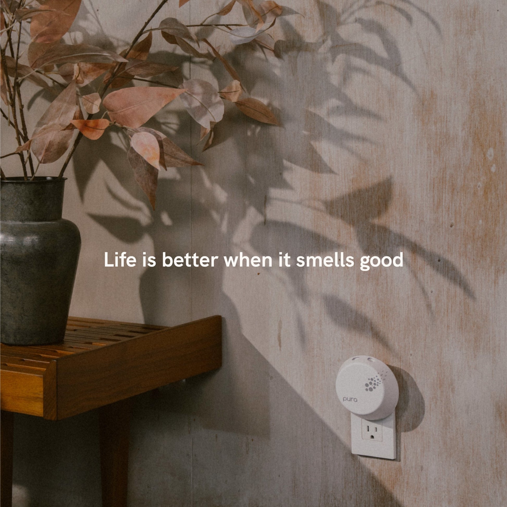 Pura smart home fragrance diffuser plugged into an outlet on a wooden surface with shadow of leaves and text 'Life is better when it smells good'