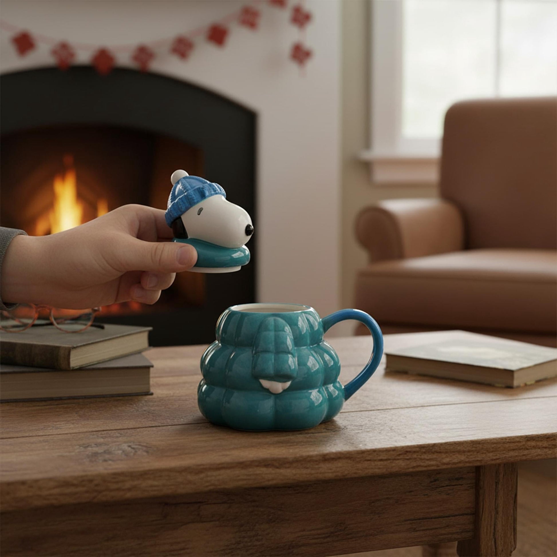 Hand holding the Snoopy lid next to the ceramic mug on a wooden table in a cozy room with fireplace background